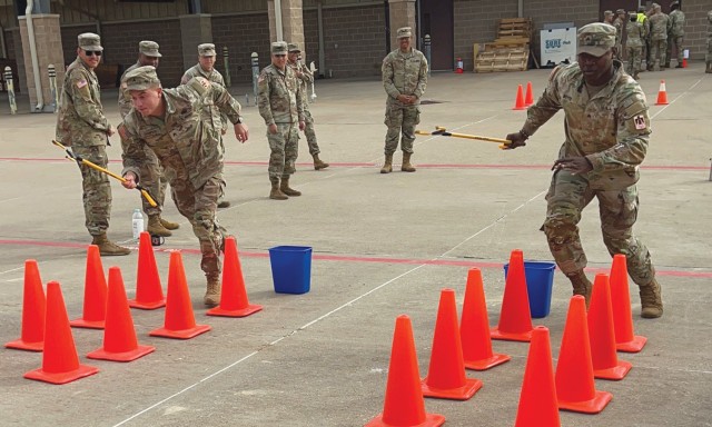 Two people dressed in operational combat pattern uniform begin to run between two separate rows of orange cones holding long grabbers.