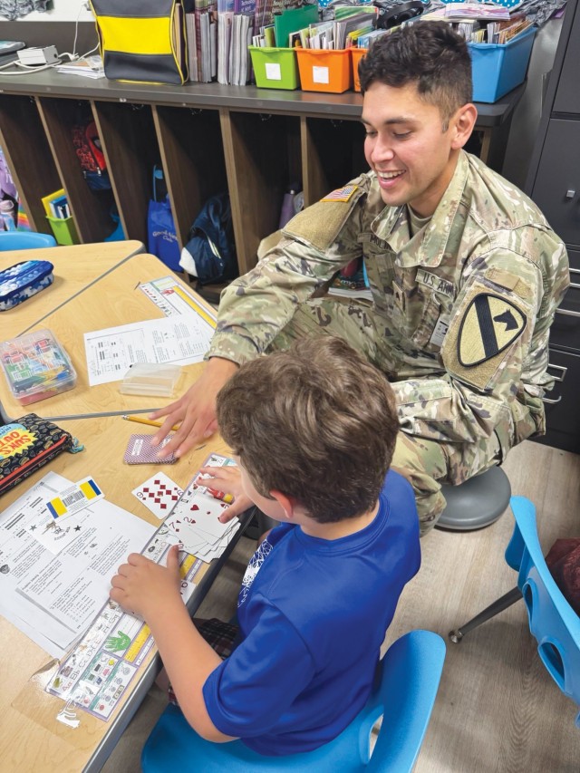 A man sits down next to a young boy, both looking at playing cards on a table in front of them.