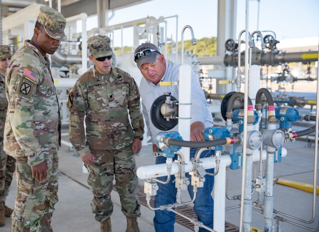 Three men stand in front of a mechanism, looking at it as the far-right man point to it. A woman stands behind them looking as well.