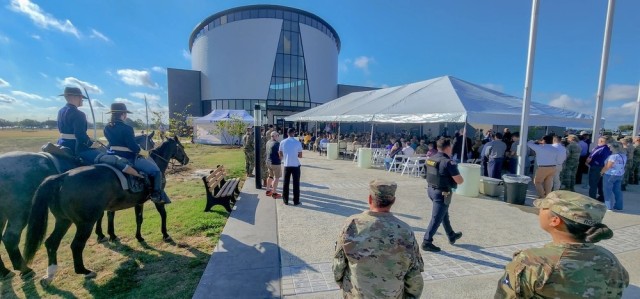 Tens of people sit and stand in front of a large, circular building, their bodies oriented toward the building.