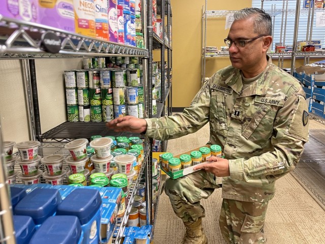A man bends on one knee in front of shelves filled with prepackaged food with a short box with rows of jars of baby food propped on his knee.