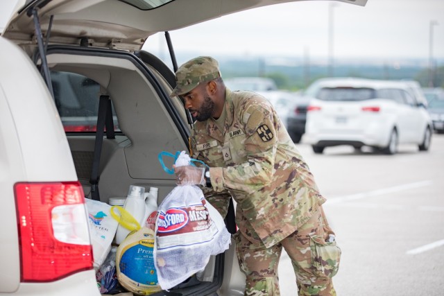 A man dress in an Army Combat Pattern uniform places a frozen turkey and firewood in a white bag in the trunk of a vehicle.