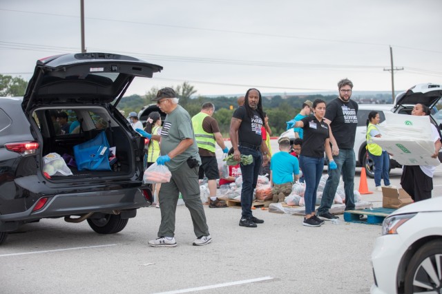 An older man moves toward an open car trunk holding a plastic bag, as other people look on.