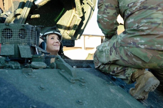 A woman smiles up from a hatch at a person squatting atop a military vehicle.