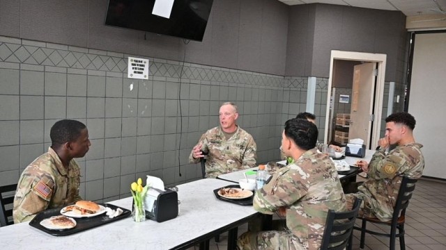 Men sit at a table with trays of food in front of them, looking at each other. One man gestures with his right hand, mouth open.