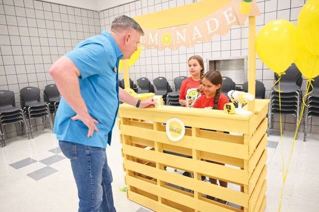 A man standing leans his left hand against a wooden stand, a cup in his hand. Behind the stand, two girls standing look on.