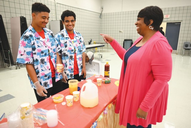 A woman and two boys standing across from each other with a red cloth-covered table smile.