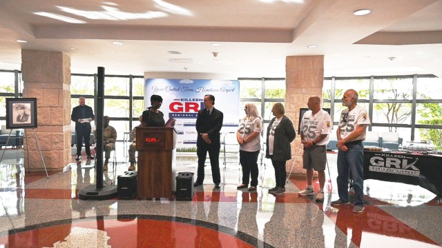 Five people stand side by side, looking at a woman standing behind a podium with a microphone on top.
