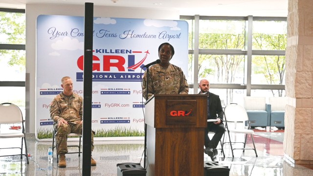 A woman stands behind a wooden podium with a microphone on a stand behind it positioned by her mouth. Behind her, two men sit in chairs looking on.