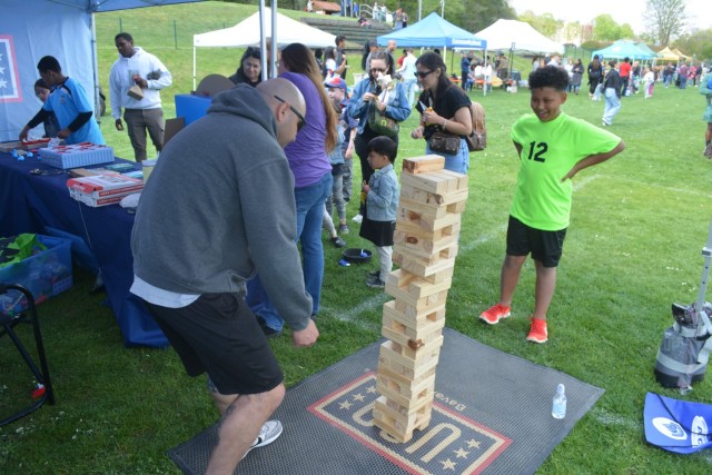 A man and a child play a somewhat large-scale game of towering blocks underneath a canopy as activities go on over a large sports field with other canopies set up thereupon.