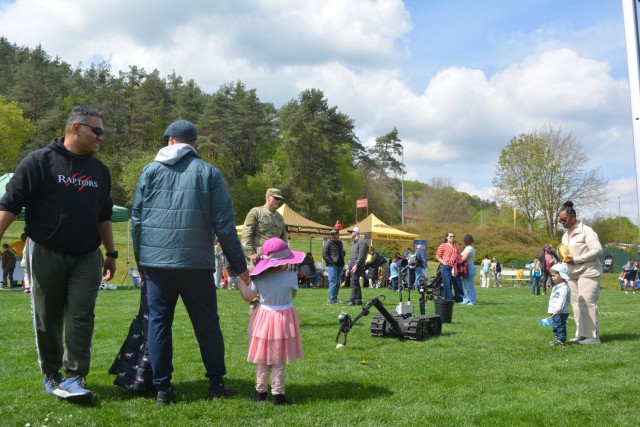 Adults and children talk to one another as a U.S. Army Soldier watches a one-armed, caterpillar-tracked vehicle pick up a baseball.