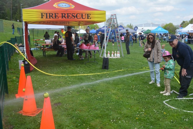 A man in blue with a ballcap helps a child, who is accompanied by her mother, knock tennis balls off traffic cones with the spray of a fire hose.