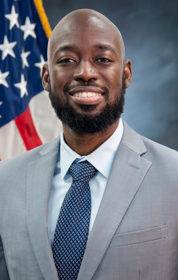 Main in gray suit and blue tie smiling in front of U.S. flag.