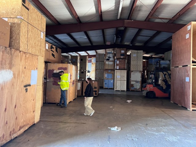 Two people walk in a large building filled with tall, stacked wooden crates.