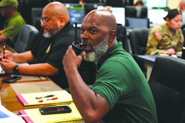 A man sitting at a table props his left elbow atop, a radio in hand close to his mouth and mouth open.