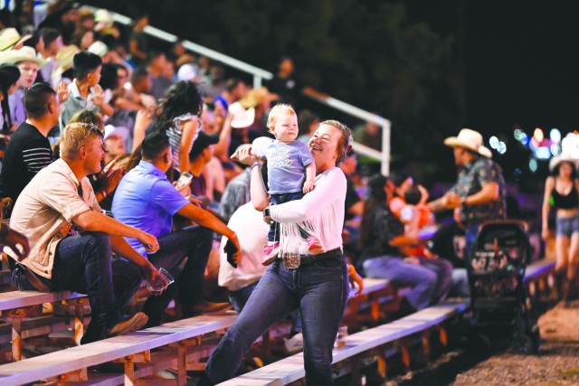 A woman smiling and standing holds an infant in her arms while looking at her.