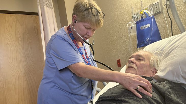 A nurse wearing stethoscope listens to the heartbeat of main lying in a hospital bed.