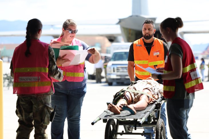 Emergency responders in bright vests coordinate around a stretcher with a covered figure. A large aircraft and vehicles are visible in the background.