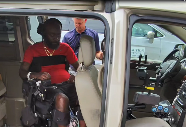 A man in a red shirt uses a wheelchair inside a van, assisted by another person outside. The vehicle appears to be adapted for accessibility.