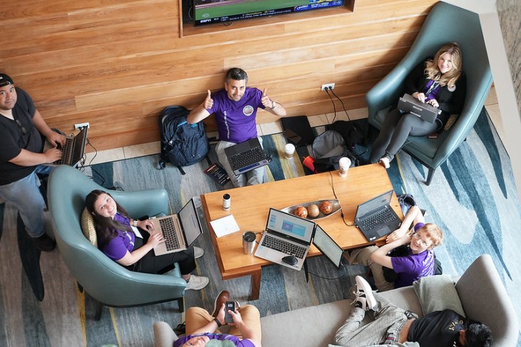 Aerial view of a group sitting in a cozy lounge with laptops, collaborating and smiling. The atmosphere is relaxed and productive, with wooden walls and soft chairs.
