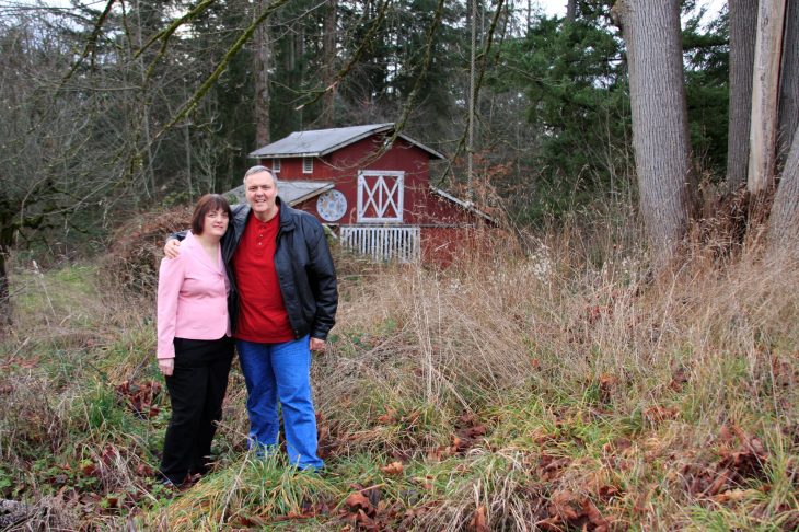 A couple stands smiling in front of a rustic red barn surrounded by tall grass and trees.