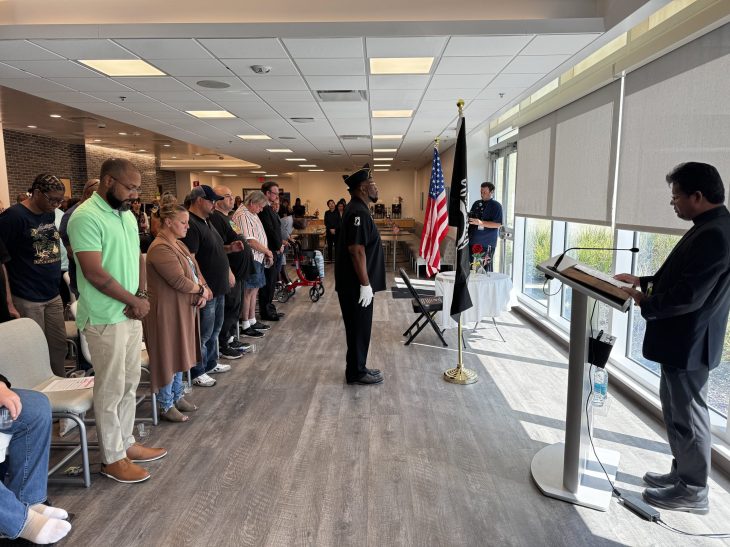 A solemn ceremony with people standing in respect near U.S. and POW/MIA flags. A man speaks at a podium.