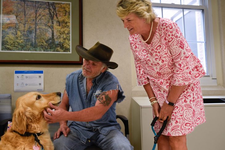 A man in a cowboy hat affectionately pets therapy dog golden retriever, while a woman in a patterned dress smiles warmly.