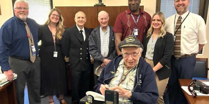 A group of eight people stands in an office, smiling warmly. An elderly man in a wheelchair, wearing a navy cap, is at the center.